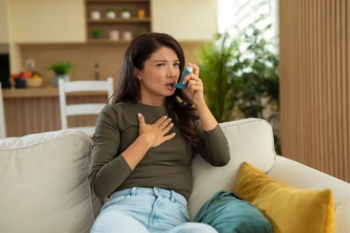 Frau sitzt auf einem Sofa und verwendet einen Asthma-Inhalator während eines Atemnot-Anfalls.