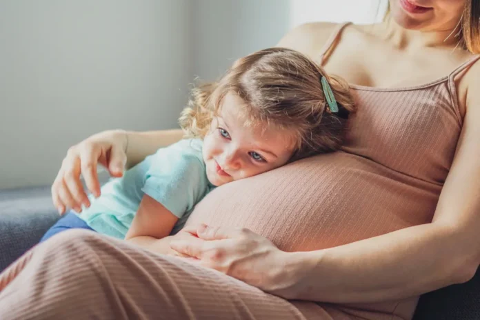 Ein kleines Kind legt den Kopf liebevoll auf den schwangeren Bauch einer Frau, die das Kind sanft hält