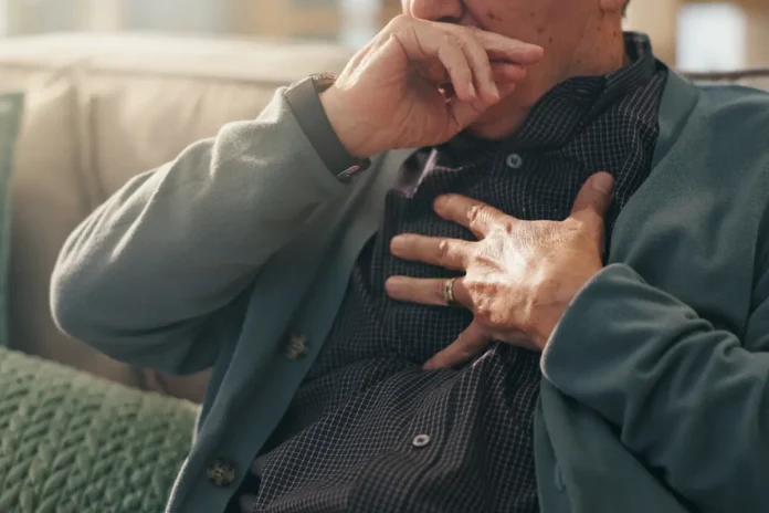 Ein älterer Mann sitzt auf dem Sofa und hält sich Brust und Mund – Anzeichen einer Atemwegserkrankung wie RSV