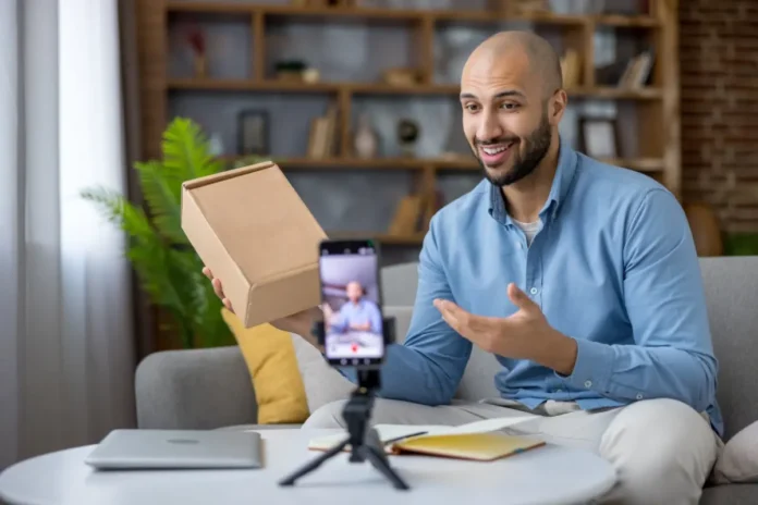Mann sitzt auf einem Sofa und nimmt mit seinem Smartphone ein Video auf, während er einen braunen Karton präsentiert. Vor ihm stehen ein Stativ mit Handy, ein Laptop und ein Notizbuch auf dem Tisch.