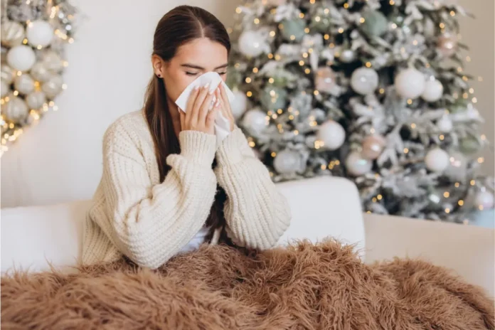 Frau sitzt eingehüllt in eine Decke auf dem Sofa und putzt sich die Nase, im Hintergrund ein beleuchteter Weihnachtsbaum