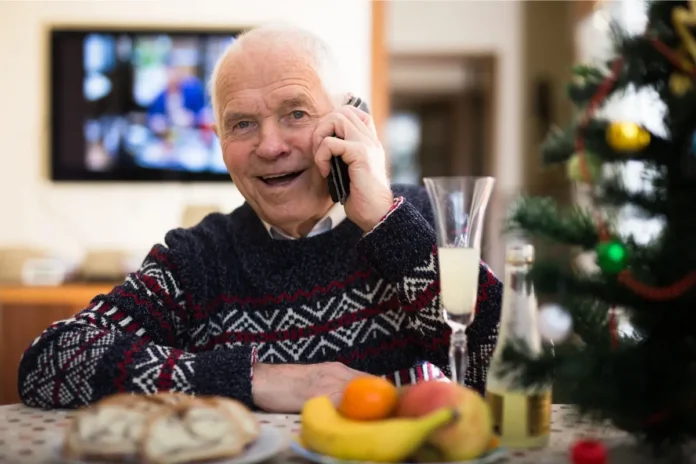 Älterer Mann sitzt an einem Tisch, telefoniert lächelnd und hält ein Glas Sekt; auf dem Tisch liegen Gebäck und Obst, im Hintergrund steht ein geschmückter Weihnachtsbaum.
