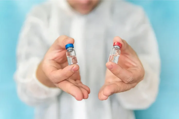Eine Person im medizinischen Schutzkittel hält zwei kleine Glasfläschchen mit klarer Flüssigkeit, eines mit blauem und eines mit rotem Deckel, in den Händen.
