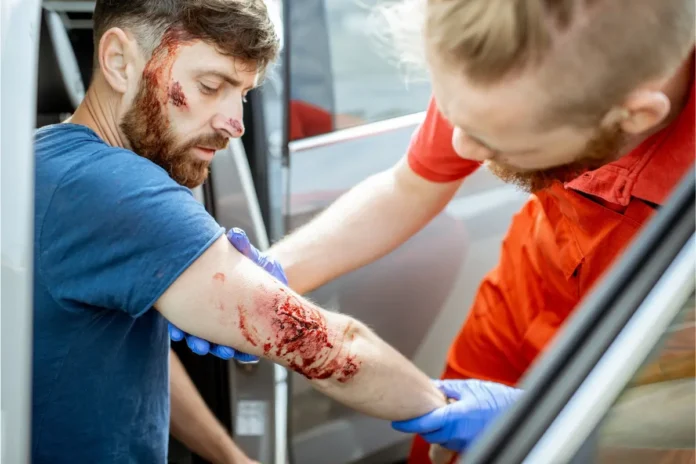Ein verletzter Mann sitzt in einem Auto, während ein Rettungssanitäter mit Handschuhen eine stark blutende Wunde an seinem Unterarm versorgt.