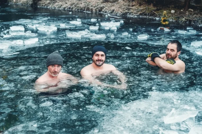 Drei Männer stehen bis zur Brust in einem eisigen Fluss mit treibenden Eisschollen und blicken in die Kamera.