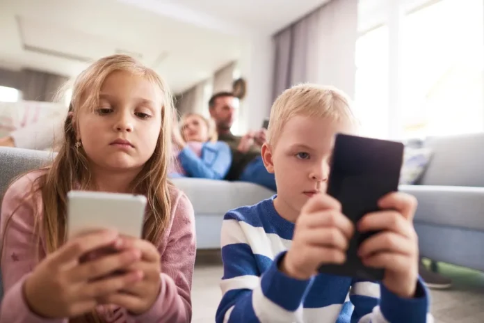 Zwei Kinder sitzen im Vordergrund und schauen konzentriert auf Smartphones, während im Hintergrund Erwachsene auf einem Sofa ebenfalls ihre Handys nutzen.
