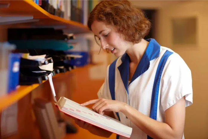 Junge Frau in weiß-blauer Arbeitskleidung liest ein Buch in einem Regalraum.