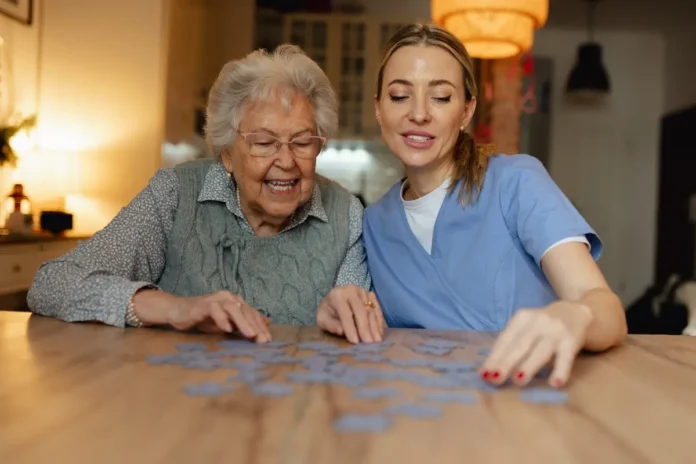 Eine ältere Frau und eine Pflegekraft sitzen gemeinsam an einem Tisch und legen ein Puzzle in einer gemütlichen Wohnküche.