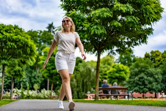 Frau in weißer Kleidung und Sonnenbrille geht auf einem Parkweg spazieren, umgeben von grünen Bäumen.