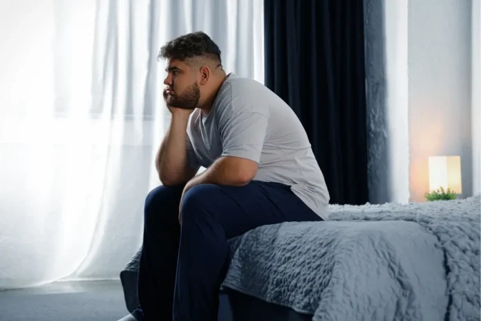 Ein nachdenklich wirkender Mann sitzt am Rand eines Bettes und blickt aus dem Fenster.