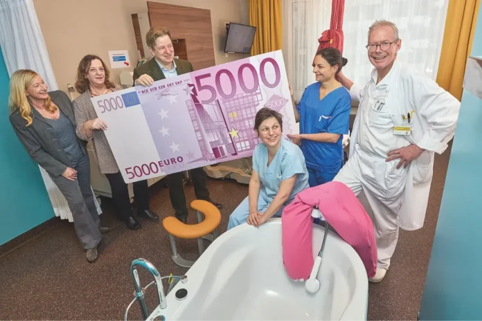 Eine Gruppe von sechs Personen steht in einem freundlich eingerichteten Krankenhauszimmer rund um eine moderne Gebärwanne. Zwei Personen halten einen überdimensionalen Scheck in Form eines 5.000-Euro-Geldscheins. Zwei Frauen in blauer Dienstkleidung und ein Mann in weißem Arztkittel lächeln in die Kamera. Im Vordergrund ist eine weiße Gebärwanne mit rosa Kissen zu sehen.