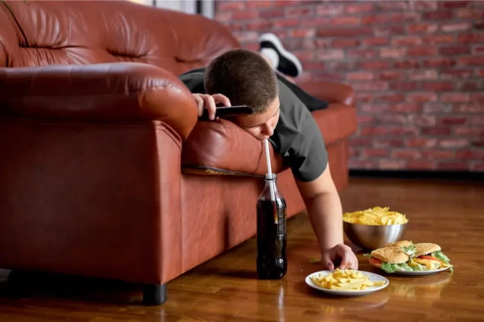 Ein Kind liegt bäuchlings auf einem braunen Ledersofa und greift nach Pommes und Burger auf dem Boden.