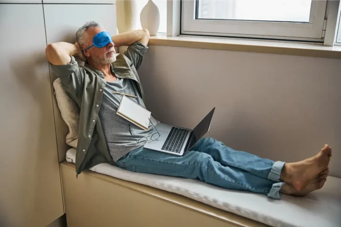Ein Mann liegt entspannt auf einer Sitzbank am Fenster, trägt eine Schlafmaske und hat einen Laptop auf dem Schoß.