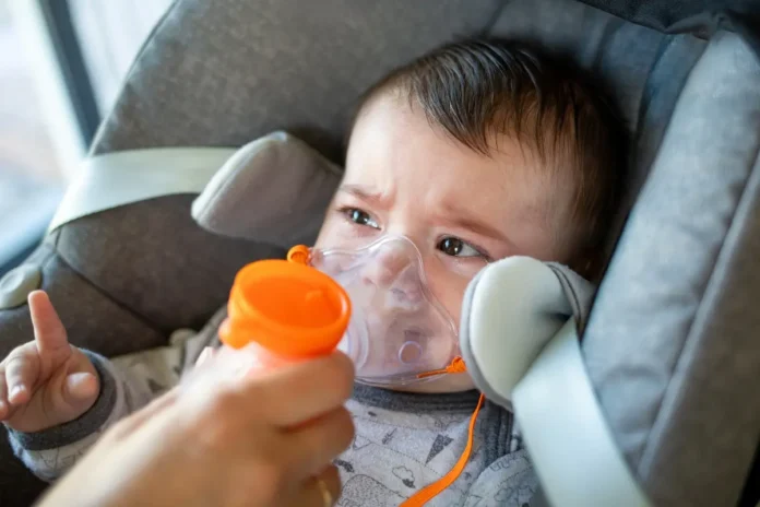 Ein Kleinkind sitzt im Autositz und trägt eine Inhalationsmaske, während eine Hand ein Inhalationsgerät hält.“