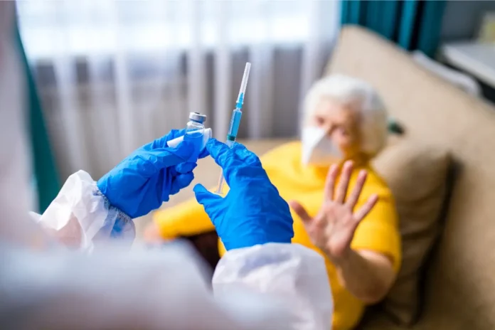 Pflegekraft mit Handschuhen hält eine Spritze, während eine ältere Person mit Maske abwehrend die Hand hebt.