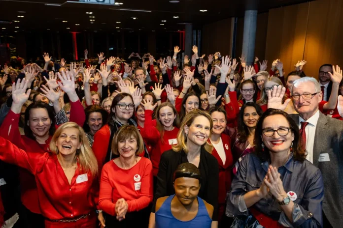 Eine große Gruppe fröhlicher Menschen – überwiegend Frauen in roten Outfits – steht dicht gedrängt zusammen, hebt die Hände und lächelt in die Kamera. Im Vordergrund sind mehrere Frauen und ein Mann gut erkennbar, einige tragen Namensschilder. Die Stimmung ist ausgelassen und positiv, im Zentrum ist eine Reanimationspuppe sichtbar.