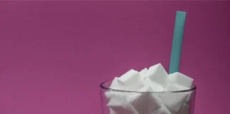 Ein Glas, gefüllt mit vielen weißen Zuckerwürfeln, in dem ein türkisfarbener Trinkhalm steckt. Der Hintergrund ist einfarbig pink.