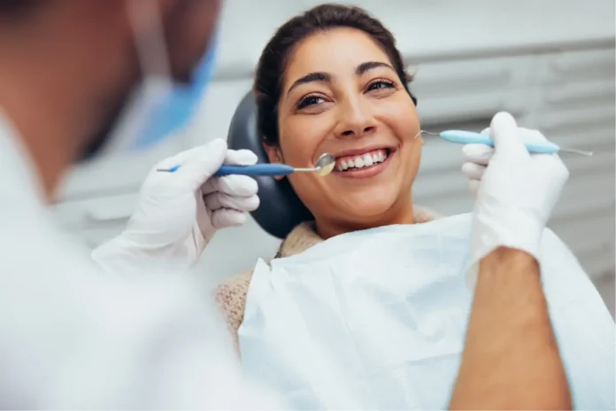 Eine lächelnde Frau sitzt beim Zahnarzt auf dem Behandlungsstuhl, während ein Zahnarzt mit Spiegel und Instrument ihre Zähne untersucht.