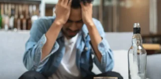 Mann sitzt mit gesenktem Kopf auf einem Sofa und hält sich die Hände an den Kopf; im Vordergrund stehen ein Glas und eine Flasche mit alkoholischem Getränk auf einem Tisch.