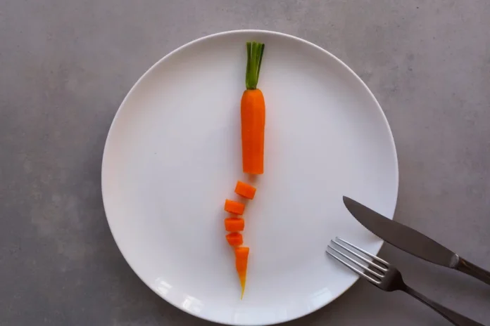 Eine teilweise in Scheiben geschnittene Karotte liegt mittig auf einem weißen Teller; rechts daneben befinden sich Messer und Gabel.