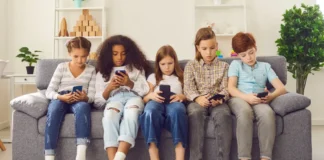 Fünf Kinder sitzen nebeneinander auf einem Sofa und schauen konzentriert auf ihre Smartphones in einem hellen Wohnzimmer.