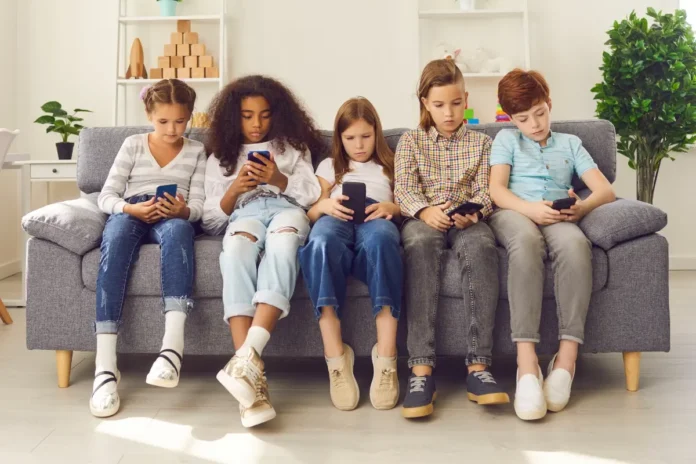 Fünf Kinder sitzen nebeneinander auf einem Sofa und schauen konzentriert auf ihre Smartphones in einem hellen Wohnzimmer.