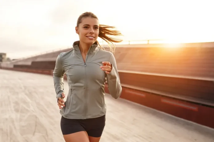 Eine sportliche Frau joggt bei Sonnenuntergang auf einem Holzsteg und trägt eine leichte Laufjacke und kurze Sporthose.