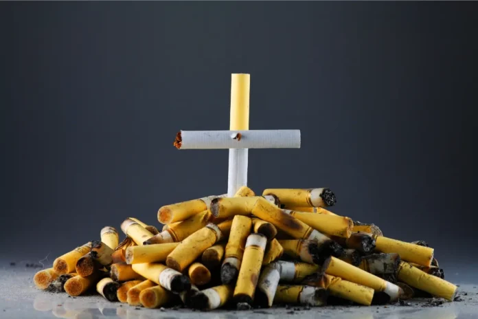 Ein Kreuz aus Zigaretten steht auf einem Haufen aus Zigarettenstummeln vor dunklem Hintergrund.