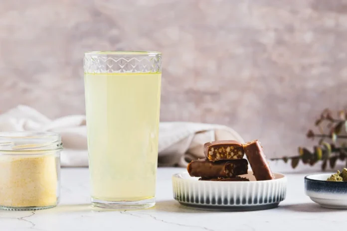 Ein hohes Glas mit hellgelbem Getränk steht auf einem hellen Tisch neben einem kleinen Teller mit Schokoriegeln.