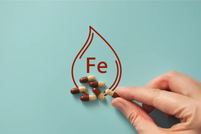 Hand hält eine Kapsel mit kleinen Kügelchen, daneben liegen mehrere ähnliche Kapseln. Im Hintergrund ist eine stilisierte Blutstropfenform mit der chemischen Abkürzung „Fe“ (für Eisen) zu sehen, auf hellblauem Hintergrund.