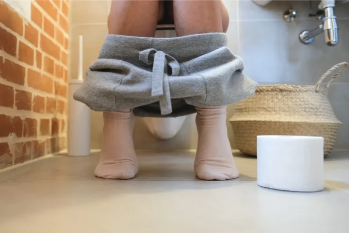 Person sitzt auf einer Toilette mit heruntergezogener grauer Jogginghose; eine Toilettenpapierrolle steht auf dem Badezimmerboden daneben.