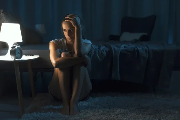 Frau sitzt nachts neben einem Bett auf dem Boden, von einer Nachttischlampe beleuchtet, und wirkt nachdenklich oder traurig, weil sie nicht schlafen kann.