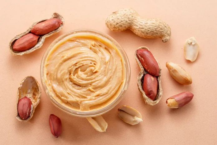 Schale mit cremiger Erdnussbutter, umgeben von ganzen und geöffneten Erdnüssen auf hellem Hintergrund.