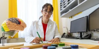 Ärztin sitzt am Schreibtisch, schreibt Notizen; vor ihr ein neuropsychologisches Testbrett mit farbigen Markern und ein Gehirnmodell.