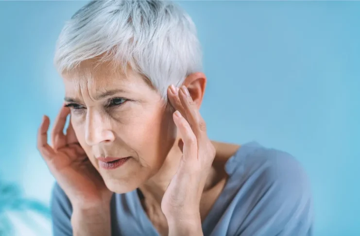 ltere Frau mit kurzen grauen Haaren hält sich konzentriert die Schläfen, möglicherweise wegen Ohrgeräuschen oder Kopfschmerzen.