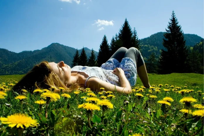 Frau liegt entspannt auf einer Wiese voller gelber Löwenzahnblüten, im Hintergrund Berge, Tannen und blauer Himmel.