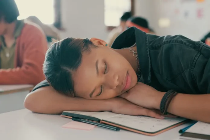 Schülerin schläft mit dem Kopf auf ihren Armen über einem offenen Notizbuch in einem Klassenzimmer.