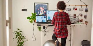 Person arbeitet an einem höhenverstellbaren Schreibtisch im Homeoffice und stützt einen Fuß auf einem Gymnastikball vor zwei Bildschirmen mit Diagrammen.