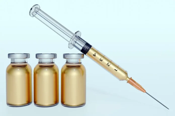 Spritze mit goldfarbener Flüssigkeit und drei kleinen Medikamentenfläschchen vor hellem Hintergrund.