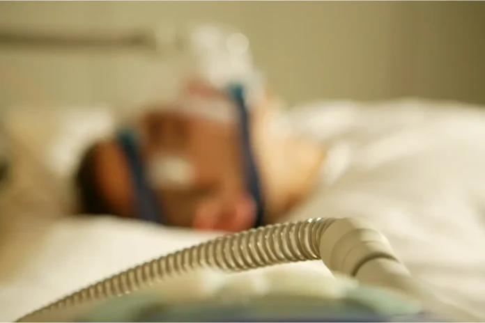 Eine Person liegt im Bett und wird mit einem Beatmungsgerät beatmet.