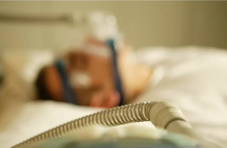 Eine Person liegt im Bett und wird mit einem Beatmungsgerät beatmet.