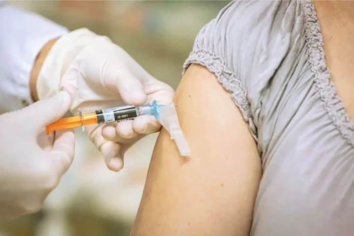 Eine medizinische Fachkraft mit Handschuhen verabreicht einer Person eine Impfung in den Oberarm mit einer Spritze.