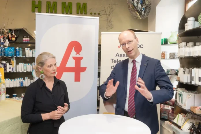 Ein Mann im Anzug und eine Frau stehen in einer Apotheke vor einem Stehtisch und sprechen miteinander; im Hintergrund sind Regale mit Apothekenprodukten und ein Banner mit einem großen roten „A“.