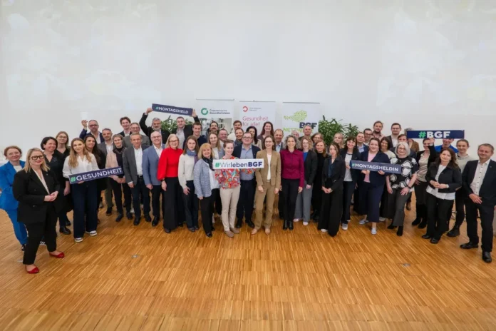 Große Gruppe von Menschen bei einer Veranstaltung zum Thema betriebliche Gesundheitsförderung, viele halten Schilder mit Hashtags wie „#WirlebenBGF“ und „#Gesundheit“.