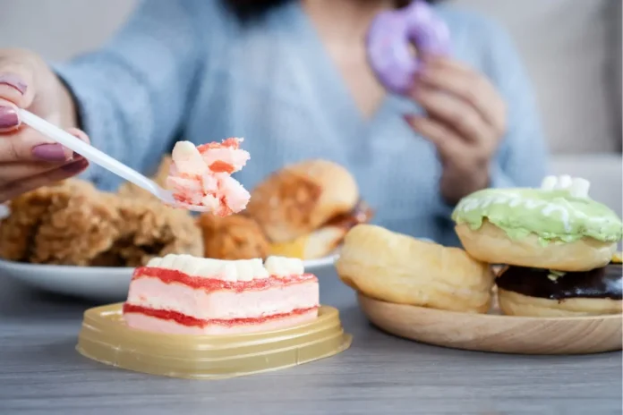 Frau sitzt vor Süßspeisen und Fast Food als Symbolbild für Essstörungen und problematisches Essverhalten