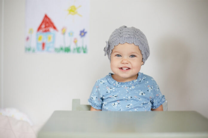 Kleinkind mit grauer Haube vor einer Kinderzeichnung