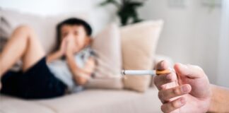 Passivrauchen in Innenräumen: Person hält eine brennende Zigarette, im Hintergrund sitzt ein Kind auf dem Sofa.