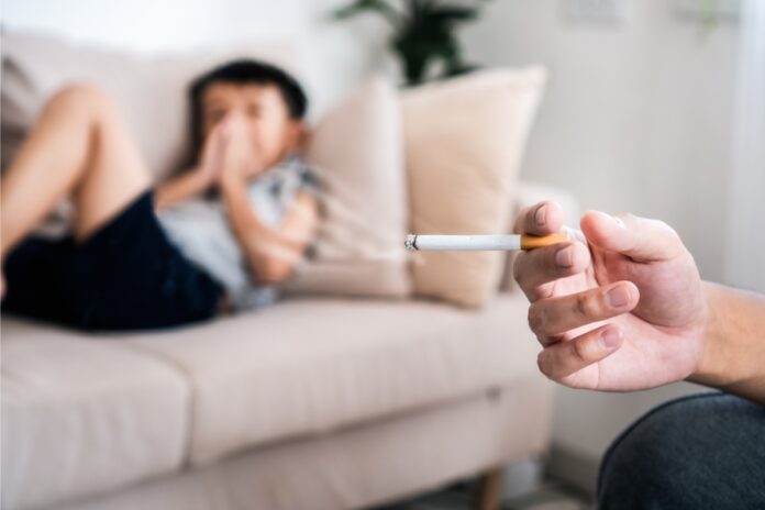 Passivrauchen in Innenräumen: Person hält eine brennende Zigarette, im Hintergrund sitzt ein Kind auf dem Sofa.