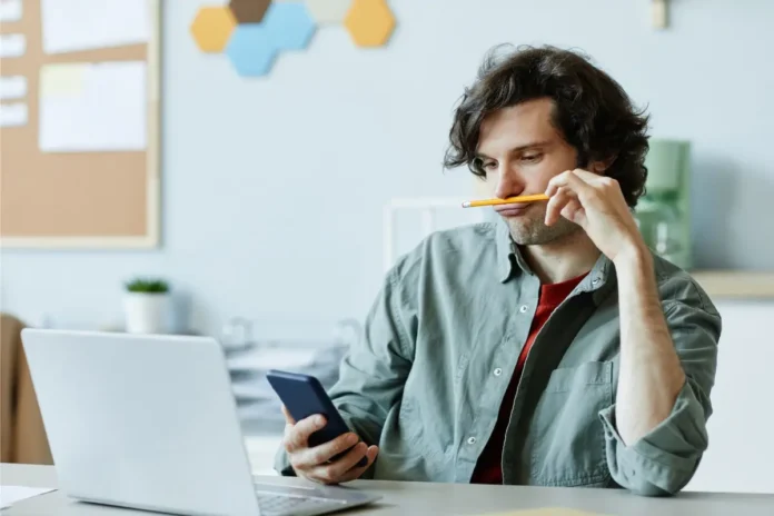 Mann blickt auf sein Smartphone, während vor ihm ein geöffneter Laptop steht.