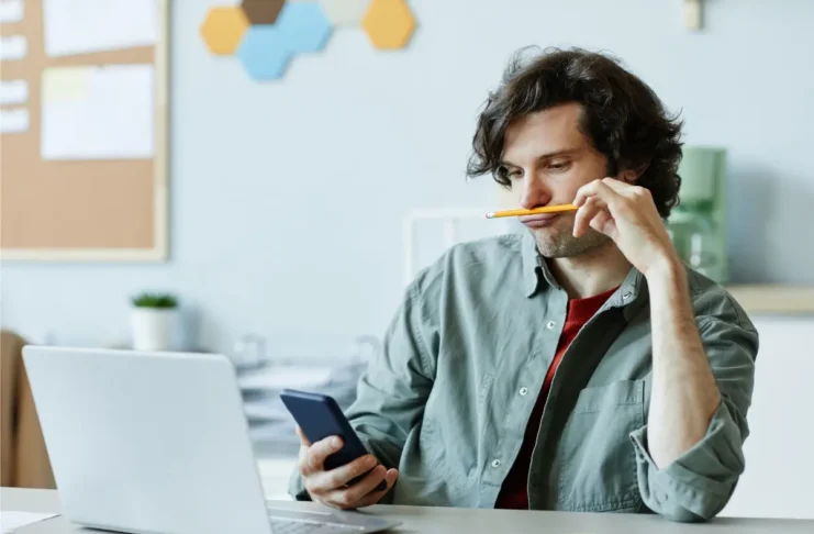 Mann blickt auf sein Smartphone, während vor ihm ein geöffneter Laptop steht.